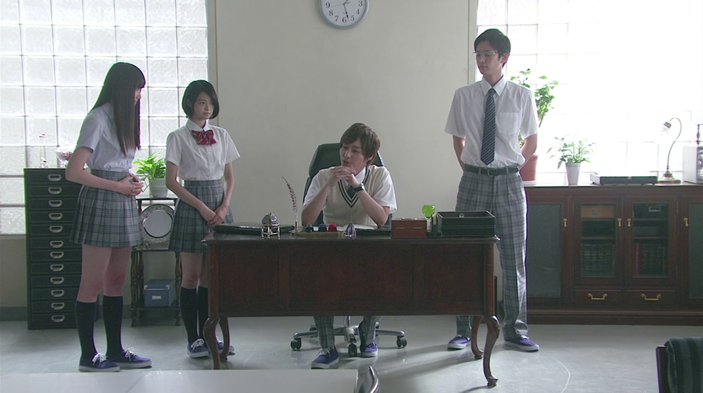 One high-school student sits impeccably dressed at an office-desk, while two female students and another male student stand around. They all emit a somewhat sinister air.